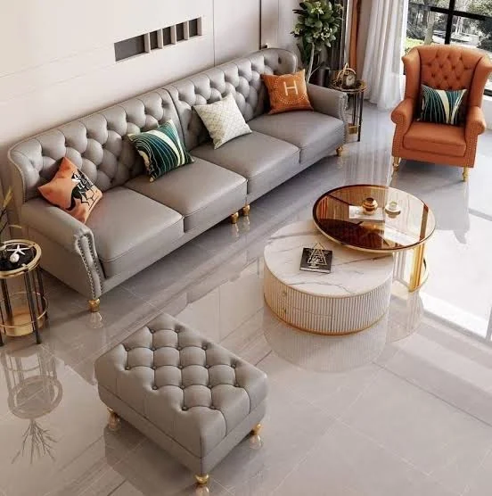 A grey tufted Chesterfield sofa and matching wingback armchair in a living room, arranged around a round brass-edged coffee table.